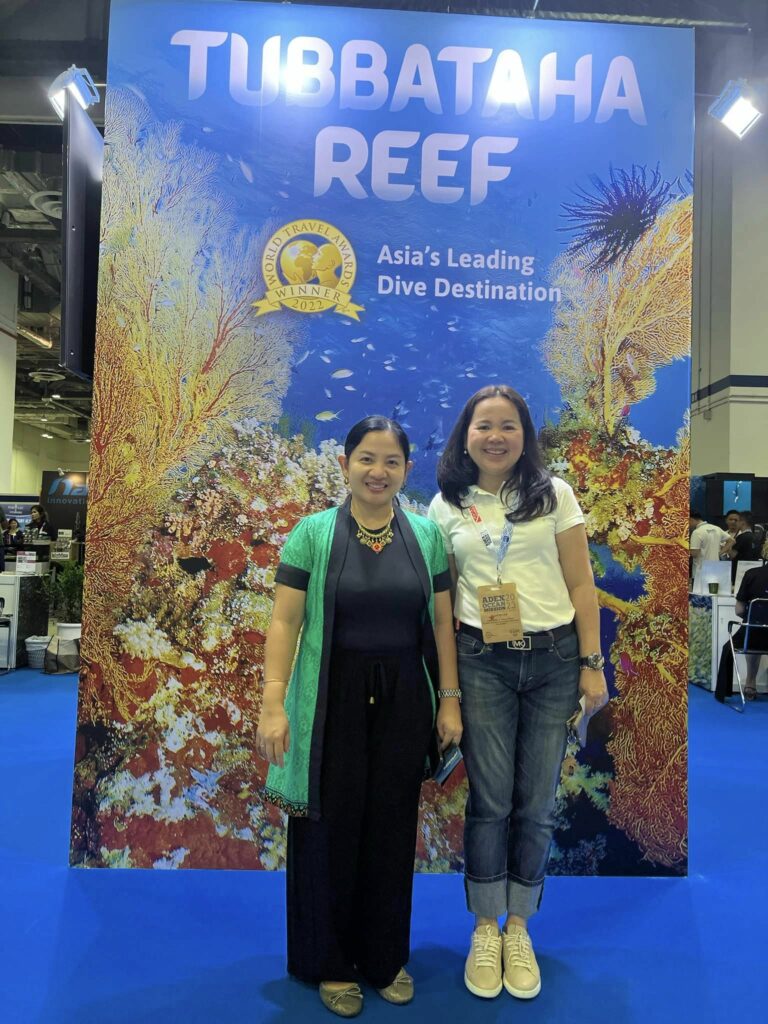 Third Secretary and Vice Consul Joy Anne Lai, Economic, Cultural and Filcom Section Head with RD Zeny Pallugna of DOT MIMAROPA pose with the Tubbataha Reef as backdrop